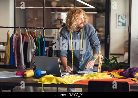 Mid-adult man designing patterns in modern fashion studio, using measuring tape and laptop Stock Photo