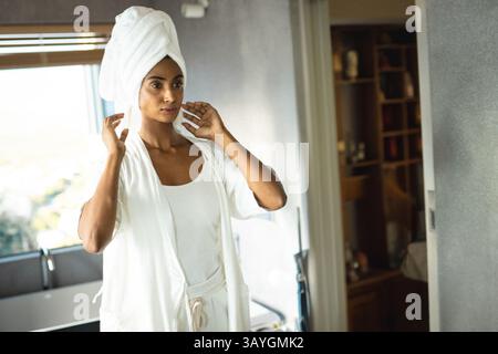 Indian Asian woman adjusting towel wrap on hair in bright bathroom, with bathrobe and blinds Stock Photo