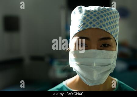 Asian female surgeon gazing at camera in hospital operating room, highlighting scrub cap and mask Stock Photo