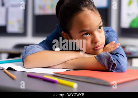 Elementary school girl resting folded arms on desk in classroom, gazing at colored pencils Stock Photo