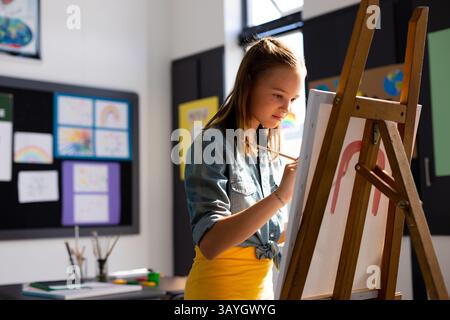 Female child painting on white canvas at wooden easel in art classroom, with paintbrush, copy space Stock Photo