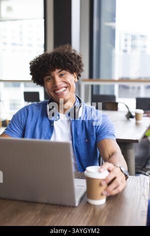 Young man working on laptop at modern cafe table, with headphones and holding paper coffee cup Stock Photo