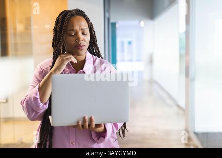 Multiracial young woman standing in modern office hallway, holding laptop and pondering ideas Stock Photo