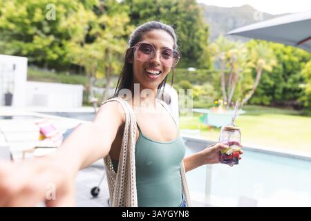 Asian woman standing at backyard pool smiling at camera, holding mason jar drink, carrying tote Stock Photo