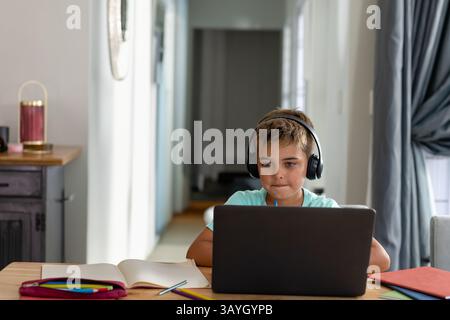 Boy sitting at wooden table in home study, using laptop with headphones and open notebook Stock Photo