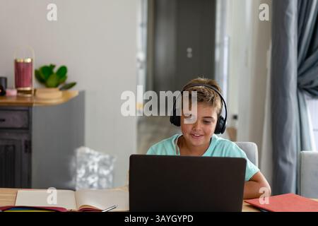 8-year-old boy sitting at wooden table in dining area, using laptop with headphones and notebook Stock Photo