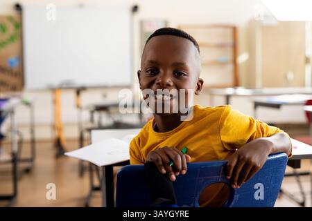 African American boy sitting backward on blue chair in classroom, holding green pencil, copy space Stock Photo