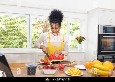 Woman slicing watermelon on cutting board on island in kitchen, with fruit bowl and tablet Stock Photo