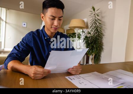 Asian man reading printed documents at wooden table in neutral home office, with potted plant Stock Photo