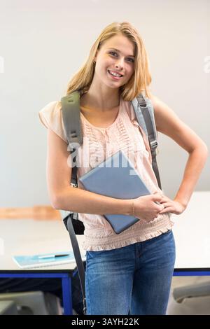 woman standing in classroom area, holding closed laptop and gray backpack, displaying notebook pen Stock Photo
