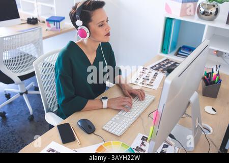 Woman typing on keyboard at desk with monitor in modern office, wearing headphones Stock Photo