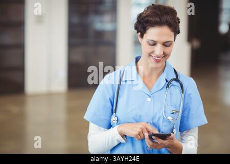Female nurse smiling while tapping smartphone in hospital hall with stethoscope, copy space Stock Photo