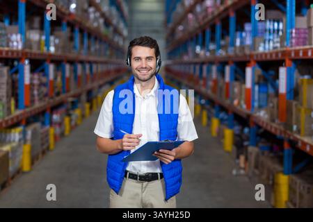 Young adult man checking inventory in warehouse aisle with clipboard pen and headset, smiling Stock Photo
