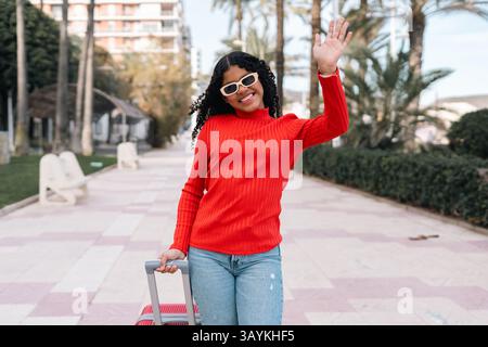 Young curly haired woman wearing sunglasses and a red sweater is pulling a trolley with one hand and waving with the other, while walking in a paved p Stock Photo