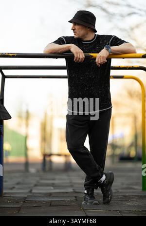 young man in black outfit and bucket hat stands casually against playground equipment. setting is park clear skies indicating vibrant day Stock Photo