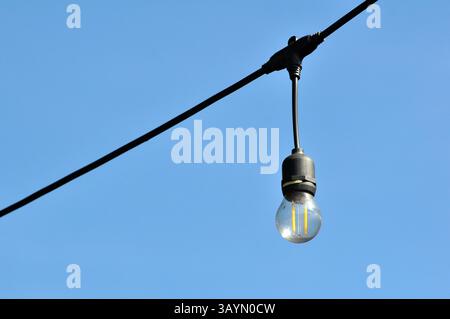 Single LED light bulb hanging from an overhead cable with a clear blue sky in the background Stock Photo