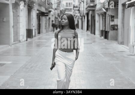 Black and white portrait of a stylish young woman walking down a city street, holding a smartphone and wearing elegant casual clothing, enjoying a lei Stock Photo