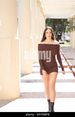 Young, stylish woman wearing a mocha mini dress and black boots, walking gracefully through a portico lined Stock Photo
