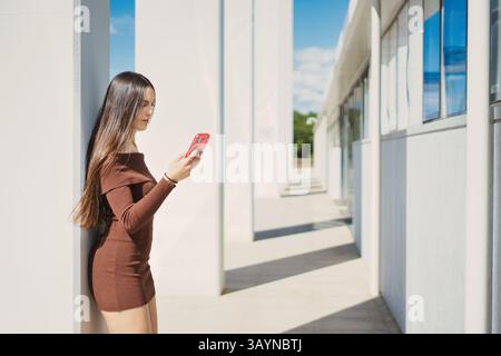 Fashionable young woman with long brown hair, wearing a mocha-colored dress, stands against a white pillar in a modern architectural space, engrossed Stock Photo