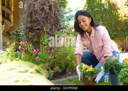 Woman kneeling in backyard garden planting yellow flowers in terracotta pot with trowel and gloves Stock Photo