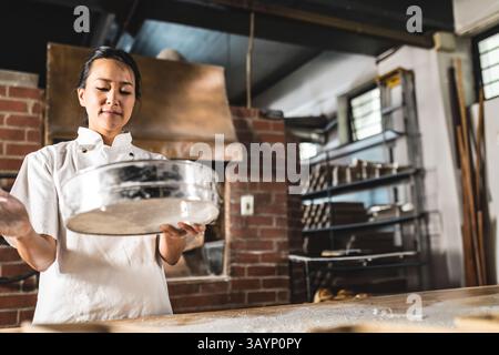 Asian woman chef sifting flour through metal sieve over countertop in bakery kitchen, copy space Stock Photo