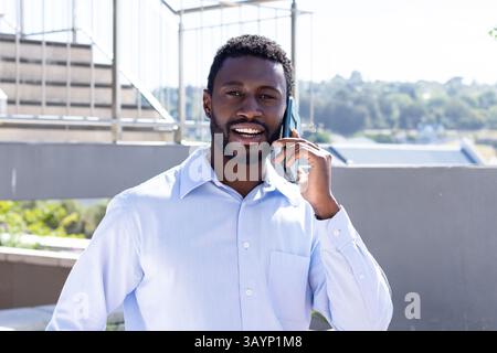 African American man standing on rooftop terrace talking on smartphone with metal railing backdrop Stock Photo