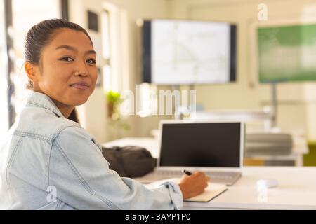 Asian female student writing in notebook at desk in classroom with laptop and screen, copy space Stock Photo