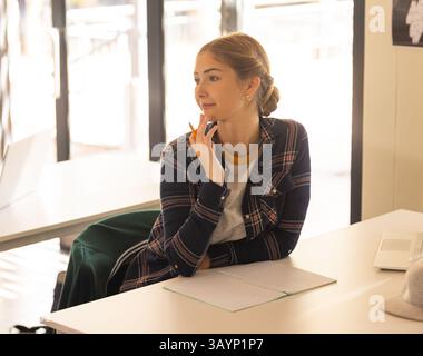 young female student holding pencil and gazing out window in study space with notebook, copy space Stock Photo