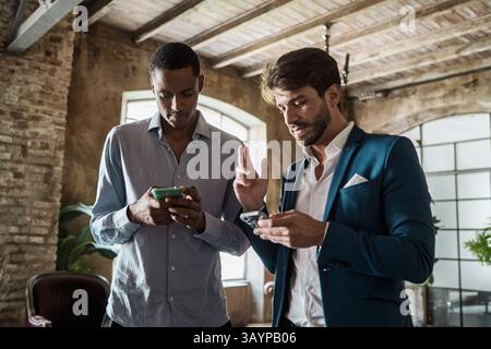 Two diverse businessmen using smartphones in a modern loft office, networking and collaborating on a project. Concept of teamwork, technology, profess Stock Photo