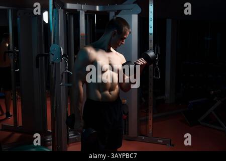 sport, bodybuilding, training and people concept - close up of young man with dumbbell flexing biceps over dark gym background Stock Photo
