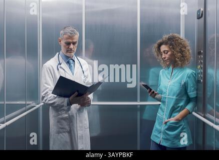 Medical staff in the hospital elevator, a doctor is checking medical records and a nurse is using her smartphone Stock Photo