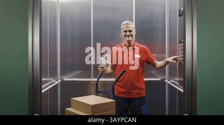 Smiling confident delivery person standing in the lift and pressing a button on the control panel, he is holding a tablet and delivering parcels using Stock Photo