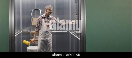 Professional painter and decorator standing in the lift and pressing a button on the control panel, he is carrying his work equipment Stock Photo