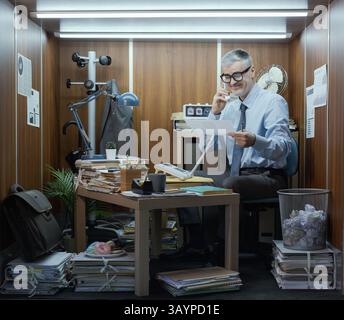 Happy office worker having a phone call in his small messy office Stock Photo