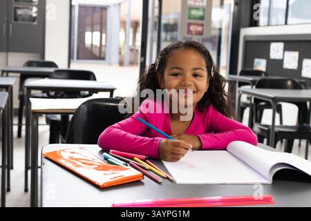Girl drawing on white paper at modern classroom desk, using colored pencils and orange notebook Stock Photo