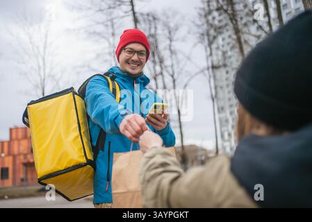 Happy male food delivery courier in blue jacket and red hat handing paper bag to customer outdoors. Contactless delivery service in urban city environment, modern lifestyle. Stock Photo