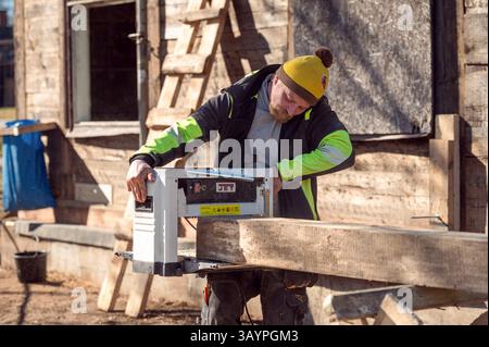Skrunda, Latvia - Mart 22, 2025: Skilled worker operates a wood planer on a beam near a wooden structure under bright sunlight. Stock Photo