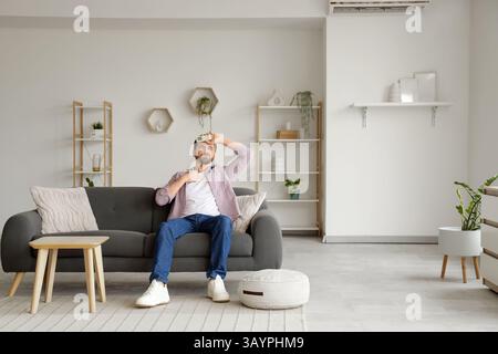 Young man with air conditioner remote control suffering from heat on sofa in living room Stock Photo