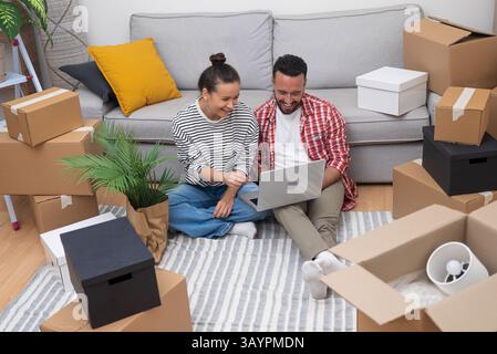 young couple, surrounded by boxes, uses laptop to shop for decor, book moving services Stock Photo