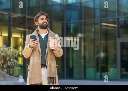 Happy Indian businessman enjoying morning coffee hot drink and using smartphone sending messages outdoors. Relaxing, taking a break. Man guy standing on urban city center street, drinking coffee to go Stock Photo