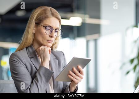 A professional businesswoman in glasses thoughtfully reviews data on a tablet in a modern office environment. Her attention is focused and engaged. Stock Photo