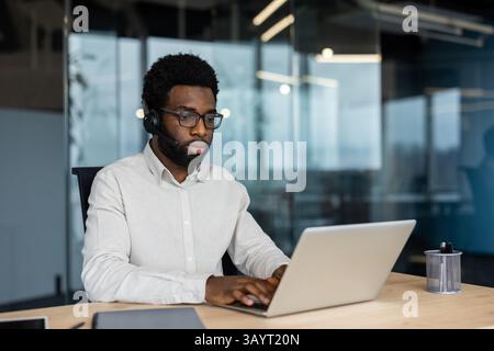 An african-american man wears a headset while working on his laptop in a modern office environment, focused on his tasks. Stock Photo