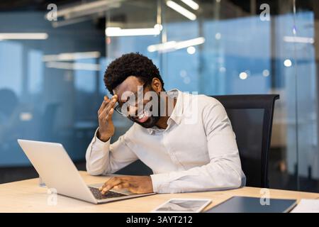 An African-American man working at his desk is shown with a stressed expression. He is experiencing a headache and appears overwhelmed while using his laptop. Stock Photo