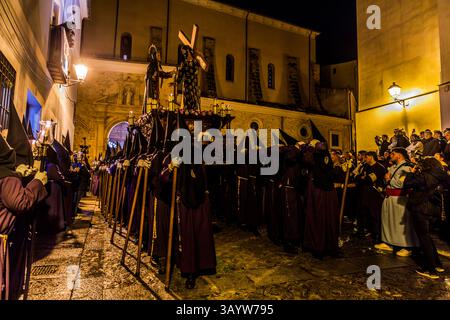 Numerous bearers in dark red robes and pointed hoods, carrying candle lanterns, process with the paso El Encuentro del Señor con la Virgen through the old town of Cuenca. Carried by the Venerable Hermandad de Nuestra Señora de la Soledad de San Agustín, the paso shows the meeting of the cross-bearing Christ with his grieving mother. Calle Alonso de Ojeda, Cuenca, Castilla-La Mancha, Spain. The paso, created in 1987 by Vicente Marín Morte, requires 32 bearers. During Semana Santa in Cuenca, 33 Hermandades take part in eleven processions through the city during Holy Week. Stock Photo