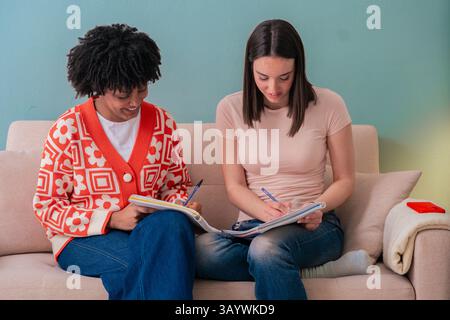 Two multiracial young women studying together on a couch, writing in notebooks with focus Stock Photo