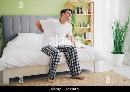 Young man experiencing discomfort in bedroom while sitting on bed during bright daytime Stock Photo