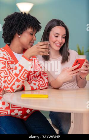 Two happy women sitting on couch smiling while looking at smartphone screen Stock Photo