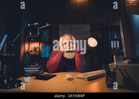 Elderly programmer works late in a cozy home office with modern technology and stylish light, focused on coding Stock Photo