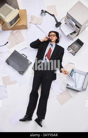 Businessman wearing in stylish formal suit lying on back holding paper and phone surrounded by office clutter. Aerial view photo. Stock Photo