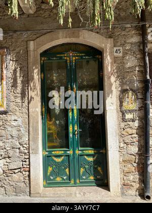 Weathered wooden door adorned with painted floral motifs, set against rustic stone wall in historic Taormina, highlighting Sicilian architectural char Stock Photo